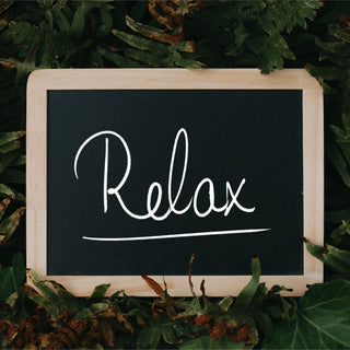 A black chalkboard with a wooden frame was white hand written text reading Relax. The chalkboard is sitting in green and brown ivy and fern leaves.
