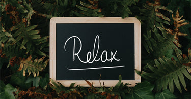 A black chalkboard with a wooden frame was white hand written text reading Relax. The chalkboard is sitting in green and brown ivy and fern leaves.
